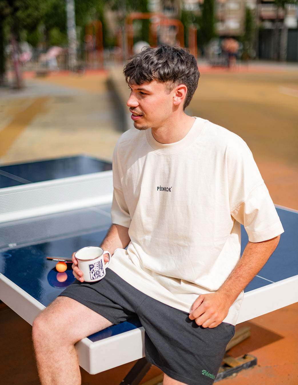 PÉENKOK' | FLORES SILVESTRES ORGANIC COTTON  SUSTAINABLE FASHION T-SHIRT WITH PÉENKOK’ PRINTED ON THE FRONT WORN BY A MAN SITTING IN A PING PONG TABLE