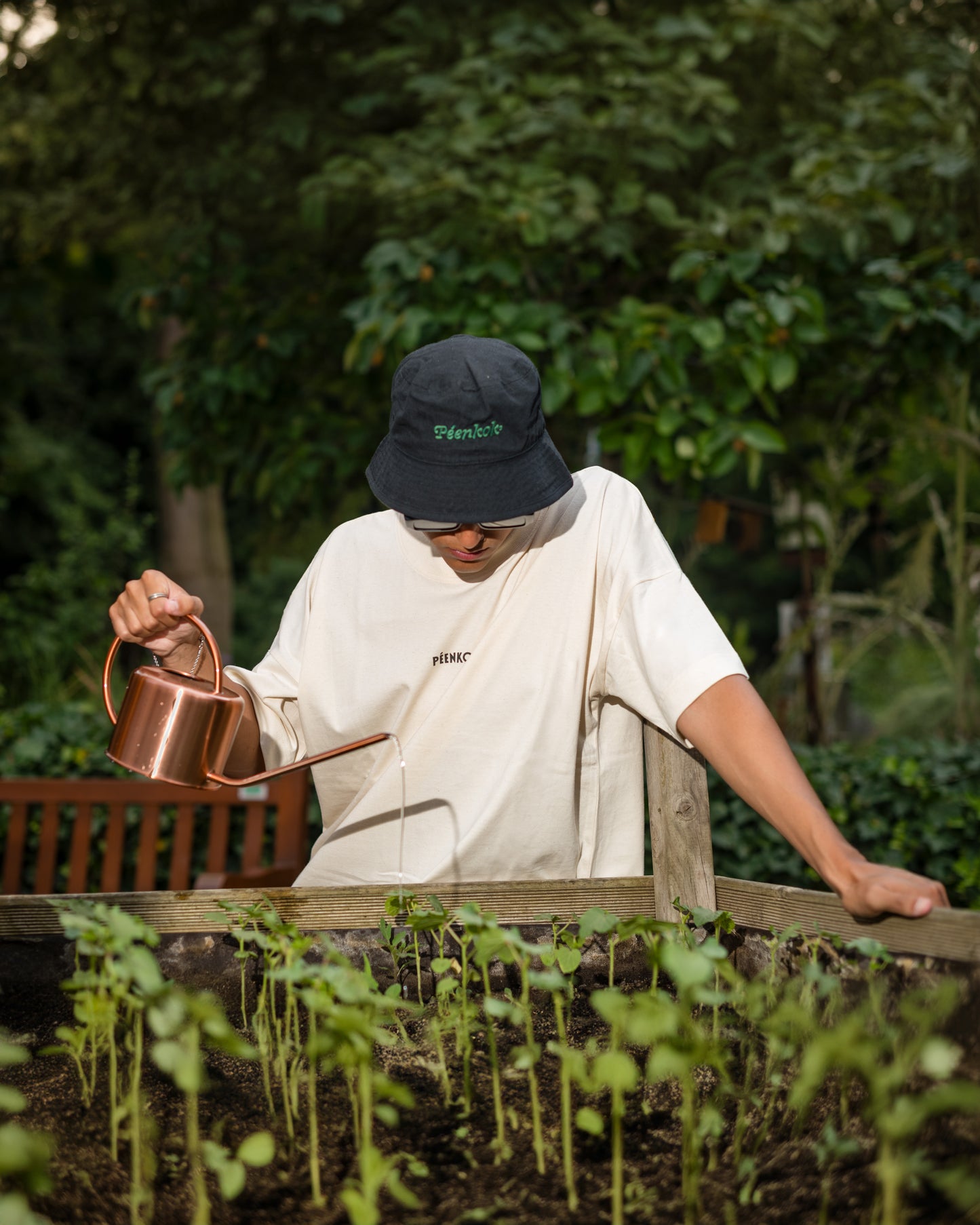 PÉENKOK' | ORGANIC COTTON SUSTAINABLE FASHION BLACK BUCKET HAT WITH ORGANIC EMBROIDERY ON THE FRONT AND ORGANIC COTTON T-SHIRT WORN BY A MAN WATERING PLANTS ON A GARDEN